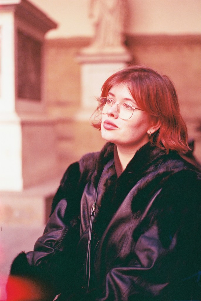 A photograph of Avery Curran, a white woman with short red hair wearing glasses and a black leather jacket with fur trim.